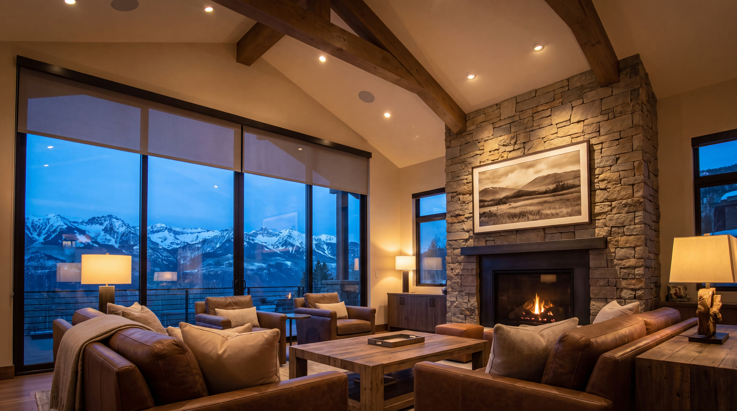 A mountain great room at dusk under exposed timber beams: a framed screen showing a black-and-white landscape set into a stacked stone chimney above a lit fire, flush ceiling speakers, motorized shades over a glass wall, and snow-covered San Juan peaks in the blue hour beyond.